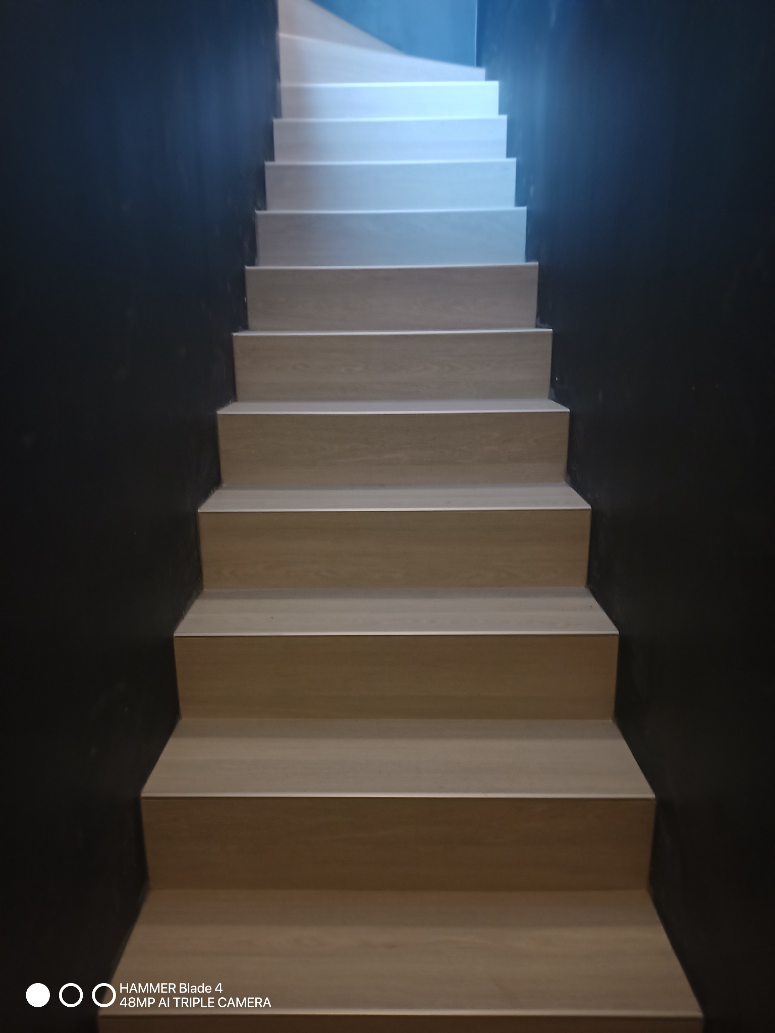 Schody wykończone panelami w odcieniach drewna, prowadzące w górę. Ściany pomalowane na ciemny kolor, kontrastujące z jasnymi stopniami. Widok z dołu.