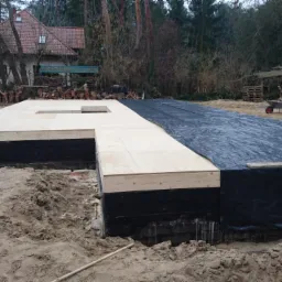 Fundament pod dom modułowy z izolacją, część przykryta czarną folią, widoczne drzewa i budynki w tle.