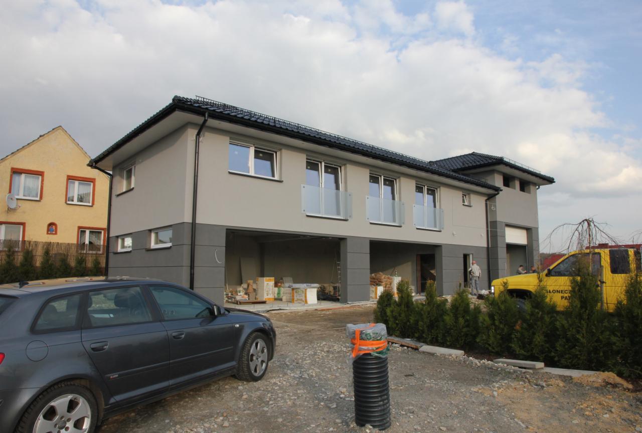 Dwukondygnacyjny budynek mieszkalno-usługowy w szarościach z balkonami, widok z zewnątrz na plac z zaparkowanym ciemnoszarym Audi i żółtą ciężarówką w tle, Racibórz-Markowice.