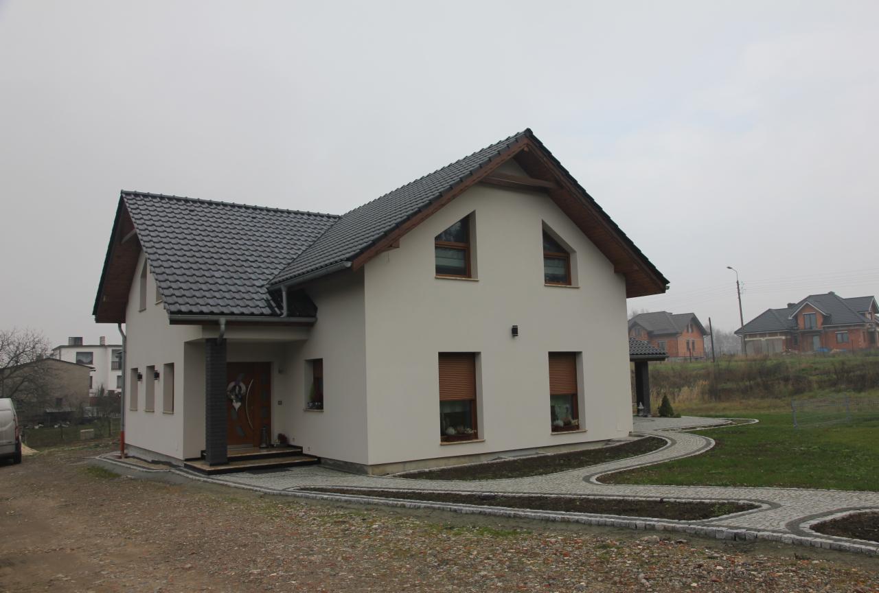 Nowoczesny dom jednorodzinny z dwuspadowym dachem pokrytym grafitową dachówką, jasną elewacją i ścieżką z kostki brukowej w Raciborzu-Brzeziu, widziany z poziomu gruntu w pochmurny dzień.