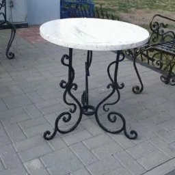 Okrągły st&oacute;ł z blatem imitującym kamień i ozdobną, kutą, czarną podstawą, stojący na szarej kostce brukowej. W tle budynki gospodarcze i zieleń.