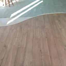 Fragment podłogi z ułożonymi panelami o drewnianym wzorze, z widocznym przejściem do płytek ceramicznych w kształcie fali i zabezpieczeniem folią malarską na górze.