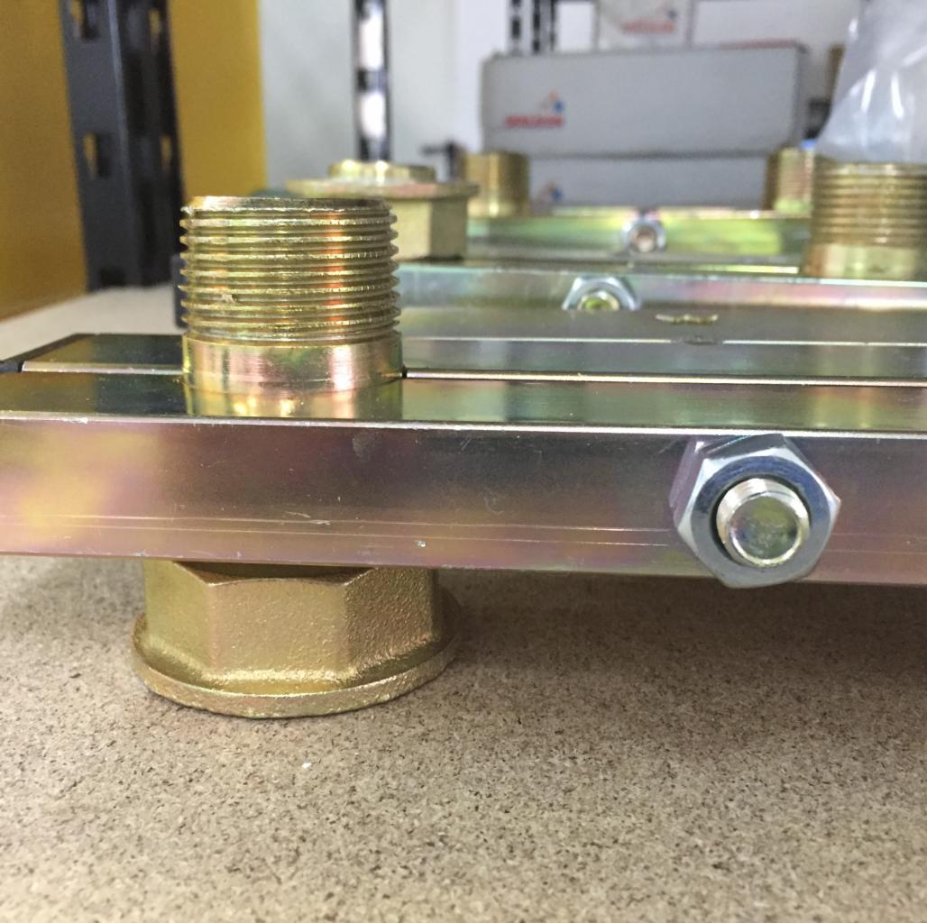 Metalowa listwa montażowa z gwintowanymi króćcami i nakrętkami, widok z bliska na szorstkiej powierzchni.