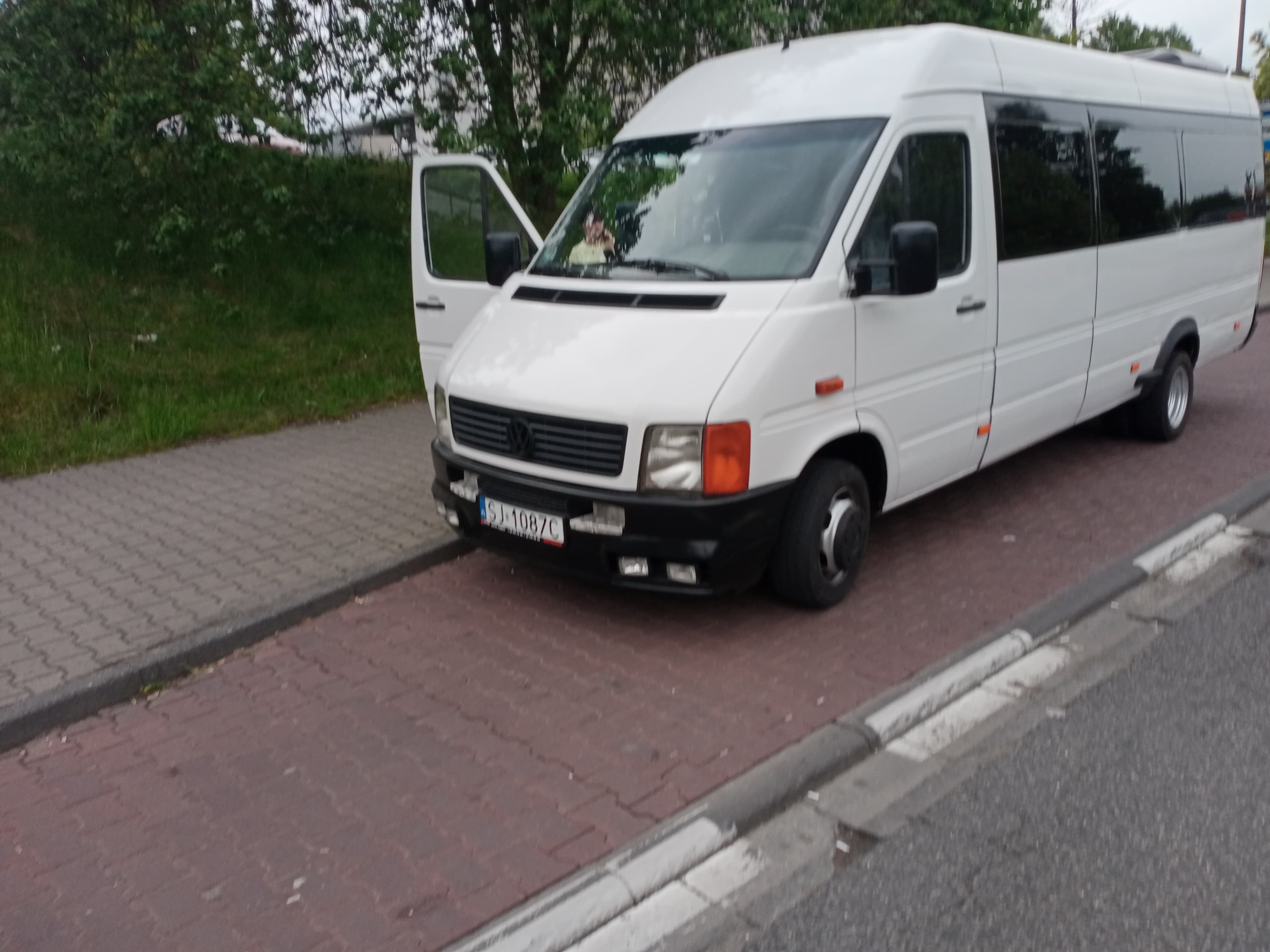 Biały bus z otwartymi drzwiami kierowcy, zaparkowany na chodniku z czerwonej kostki, widoczny numer rejestracyjny.