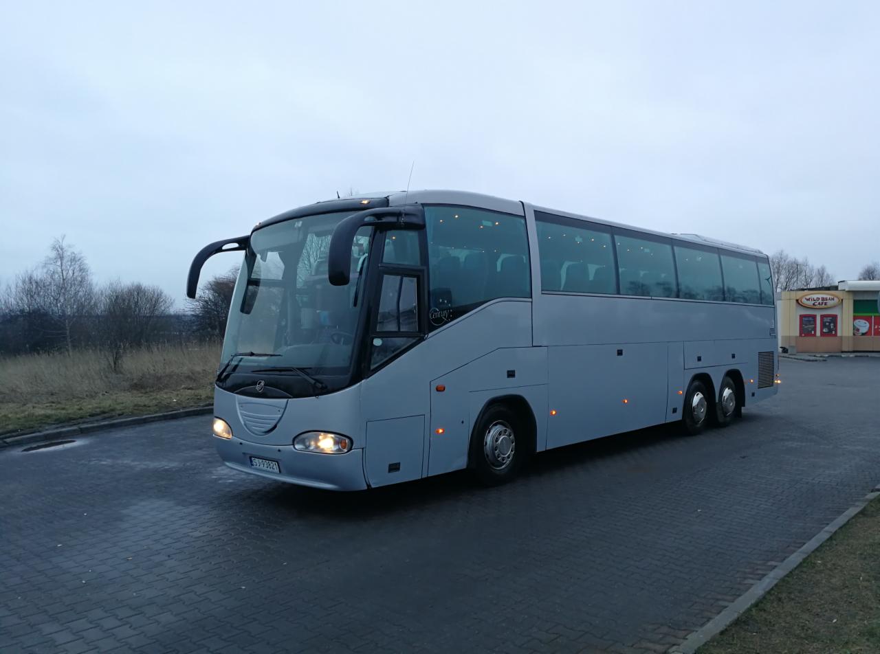 Szary autokar turystyczny w Jaworznie, zaparkowany na brukowanym placu, widoczny z przodu i boku, z włączonymi światłami pozycyjnymi, w tle budynek kawiarni.