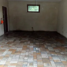 Wnętrze garażu z nową, wzorzystą posadzką z płytek imitujących drewno, surowe ściany przygotowane do wykończenia, okno w tle.