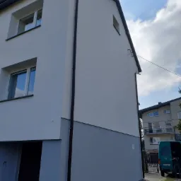 Dwukondygnacyjny dom jednorodzinny z jasną elewacją, pomalowaną na dwa odcienie szarości; widoczne okna z odbiciem nieba oraz brązowe rynny. W tle zaparkowany bus.