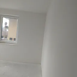 Pomalowane na biało wnętrze pokoju z oknem, widoczne ślady po pracach wykończeniowych na podłodze.