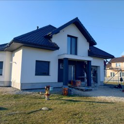 PIK-EL USŁUGI BUDOWLANE DAWID PIKUL - Dwukondygnacyjny dom jednorodzinny w trakcie malowania elewacji. Widoczne rusztowania, farby i pracownicy w kombinezonach. Na trawniku stoi ozdobna figurka.