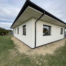 PIK-EL USŁUGI BUDOWLANE DAWID PIKUL - Nowoczesny, parterowy dom z jasną elewacją i czarnymi ramami okiennymi oraz rynnami. Widoczny fragment trawnika i osoba pracująca w oddali.