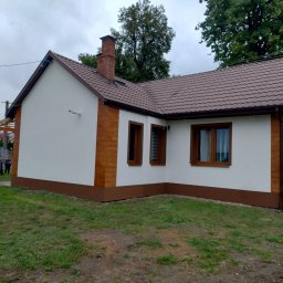 PIK-EL USŁUGI BUDOWLANE DAWID PIKUL - Biały dom z brązowym dachem i elementami dekoracyjnymi imitującymi drewno na elewacji, widoczny zieleń trawnika i częściowo samochód dostawczy firmy.