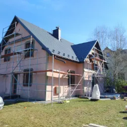 Dom jednorodzinny w trakcie ocieplania elewacji, widoczne rusztowania, częściowo otynkowane okna i pracownik na rusztowaniu, na pierwszym planie narzędzia i samochód osobowy.