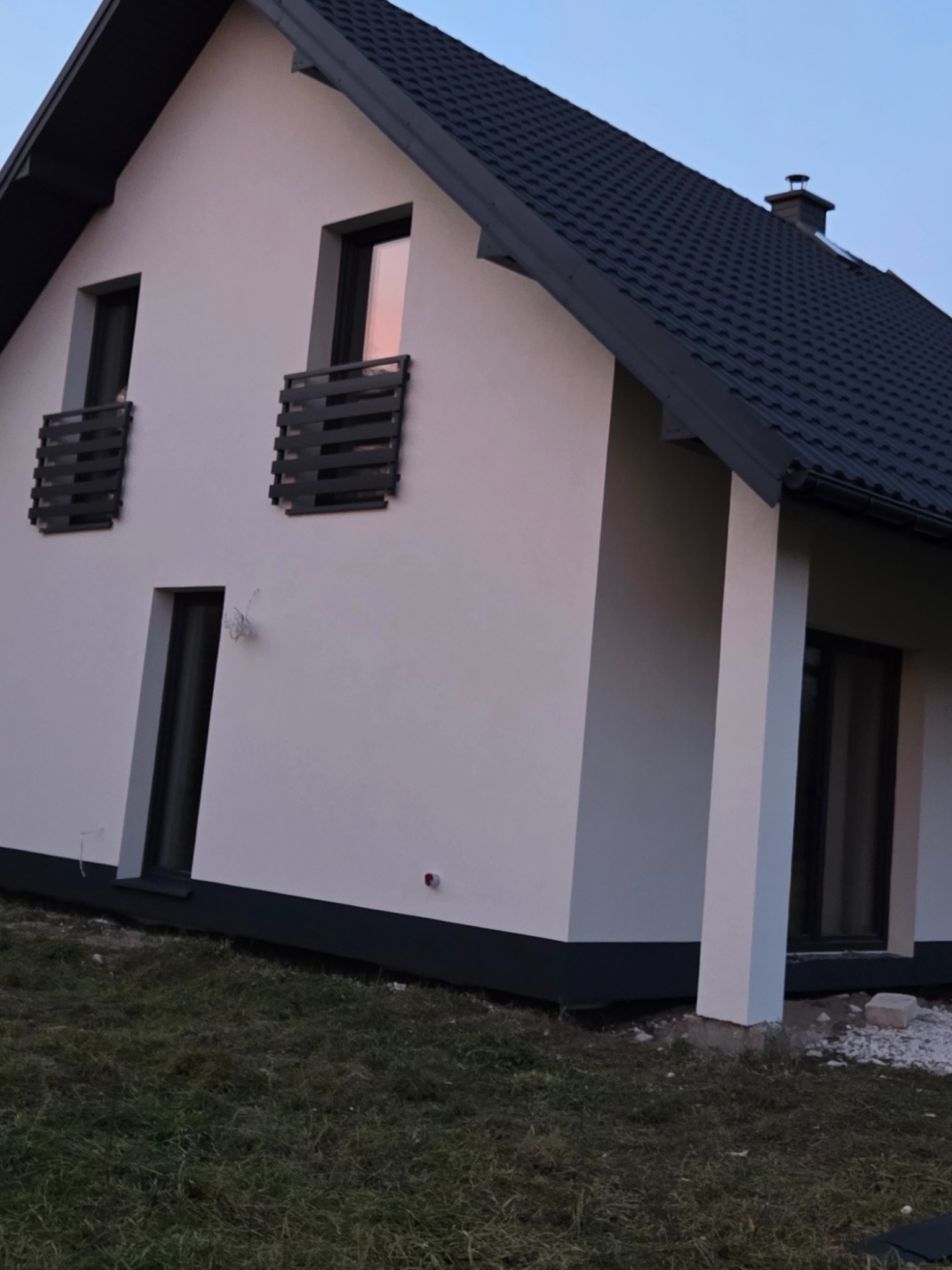 Biały dom jednorodzinny z ciemną dachówką i stolarką okienną, świeżo po malowaniu elewacji, widok z boku na tle błękitnego nieba o zmierzchu.