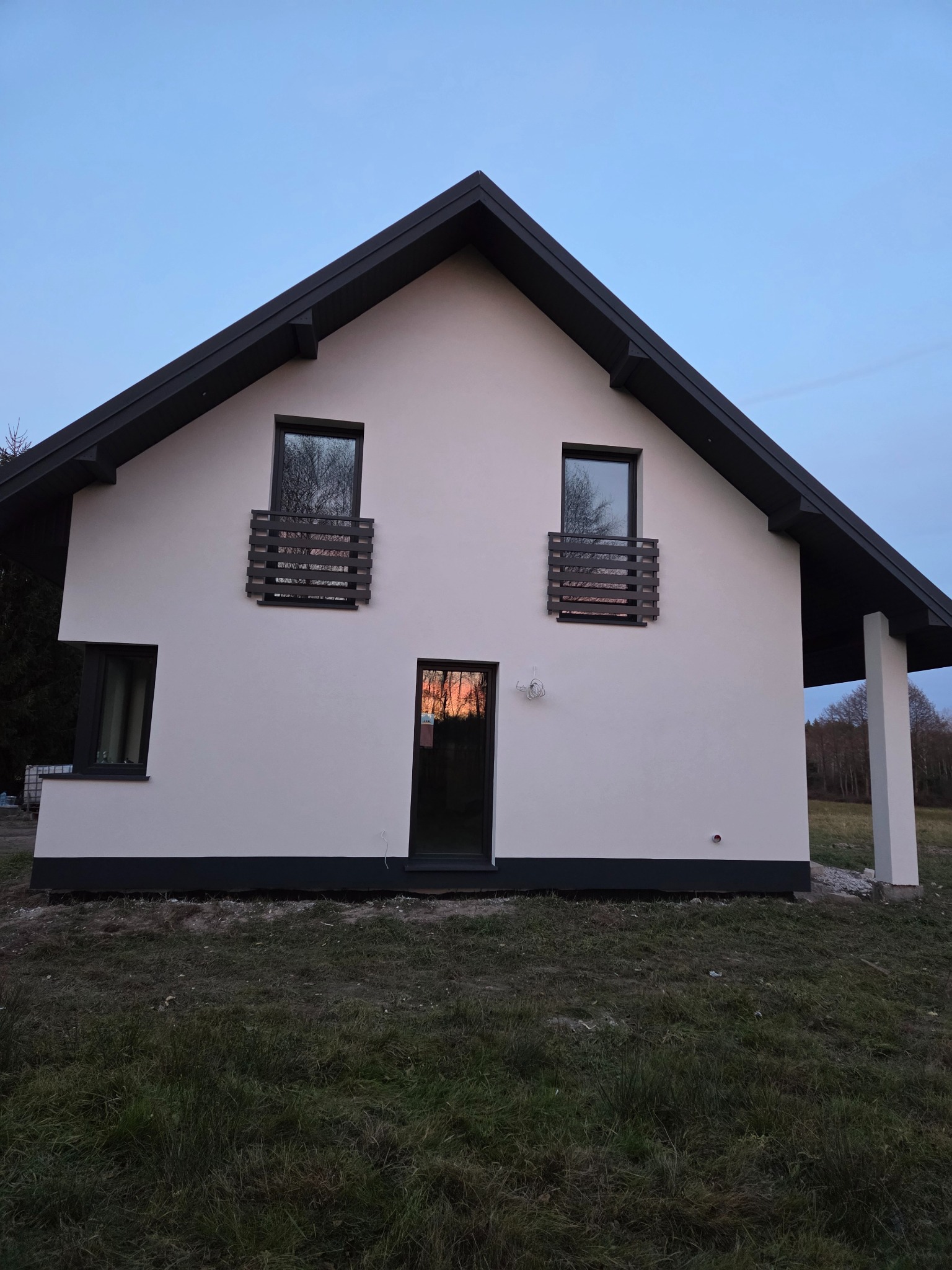 Biały dom jednorodzinny z ciemnymi oknami i dachem, widziany z perspektywy trawnika. W oknach odbija się zachód słońca, a na elewacji widoczne są drewniane elementy dekoracyjne.