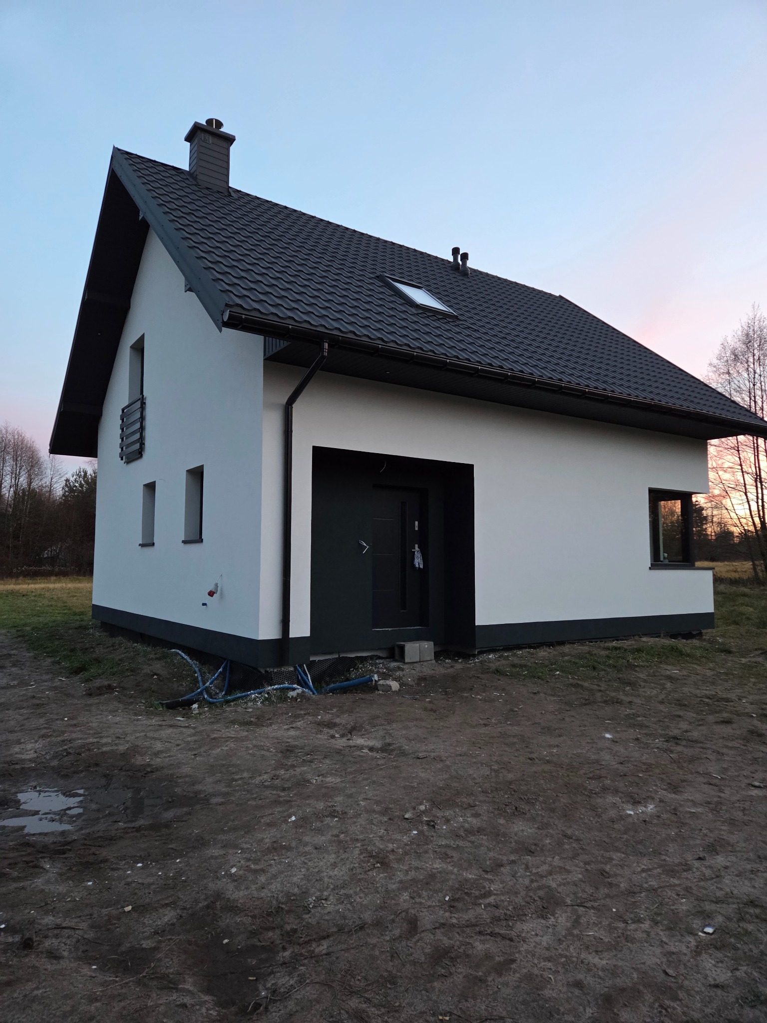 Nowoczesny dom jednorodzinny z białą elewacją i grafitowym dachem, widoczny z zewnątrz o zmierzchu. Uwagę zwraca minimalistyczny design i staranne wykończenie.