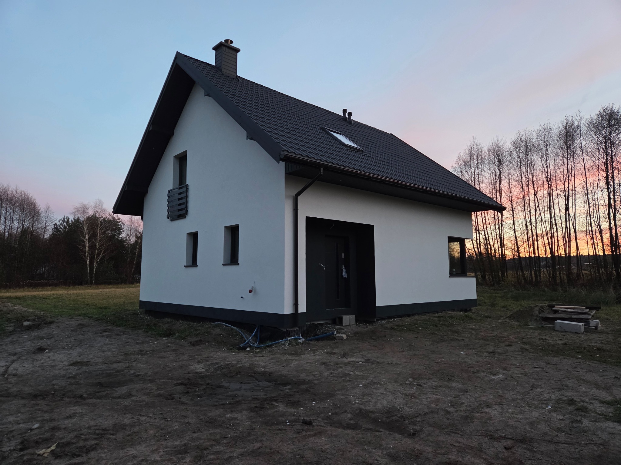 Biały dom jednorodzinny z ciemną stolarką i dachem na tle zachodzącego słońca. Widoczne elementy elewacji i otoczenie w stanie surowym.