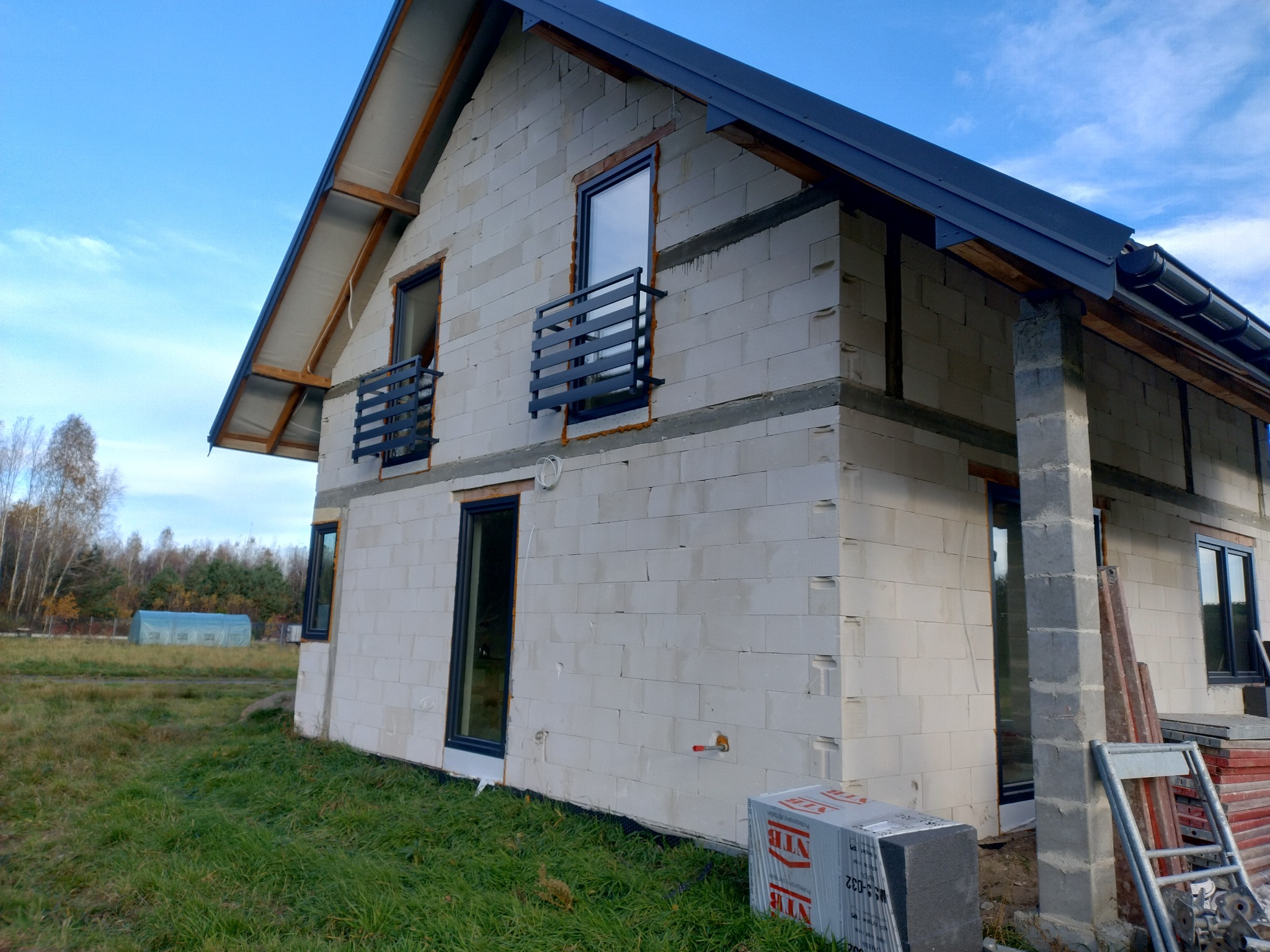 Niedokończony dom z białych bloczków, widoczne okna z czarnymi ramami i balustrady balkonowe. Na pierwszym planie trawa i materiały budowlane.