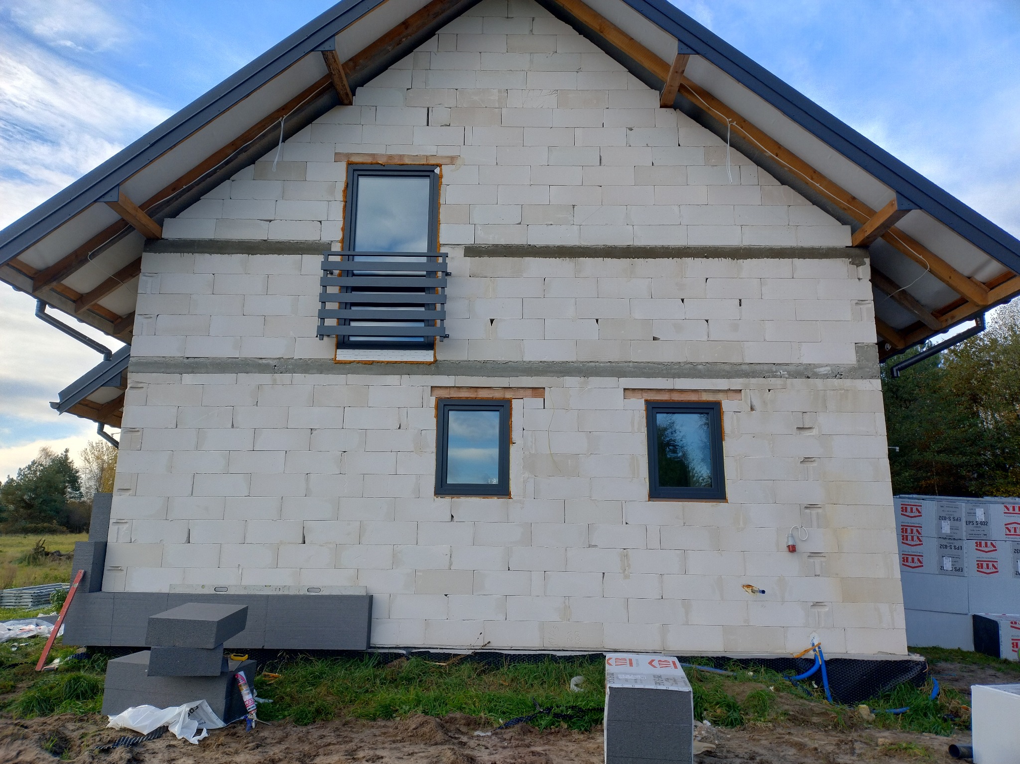 Surowy budynek z betonu komórkowego w trakcie budowy, z zamontowanymi oknami i przygotowanymi materiałami izolacyjnymi. Widoczne bloczki i ocieplenie.