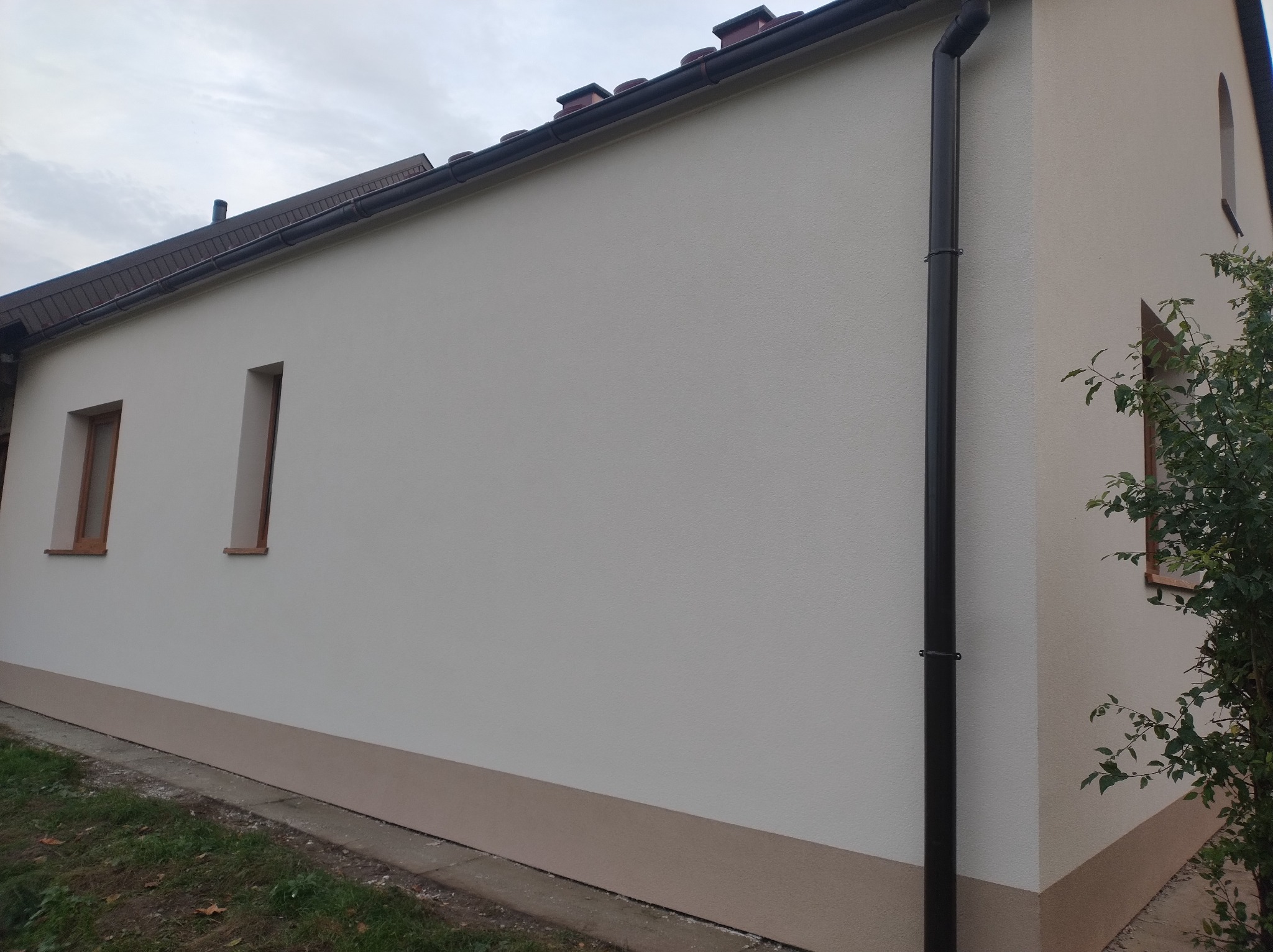 Jasna elewacja budynku jednorodzinnego z brązowymi oknami i rynną. Widoczny fragment dachu z brązową dachówką i cokół w odcieniu beżu. Świeżo pomalowana ściana.