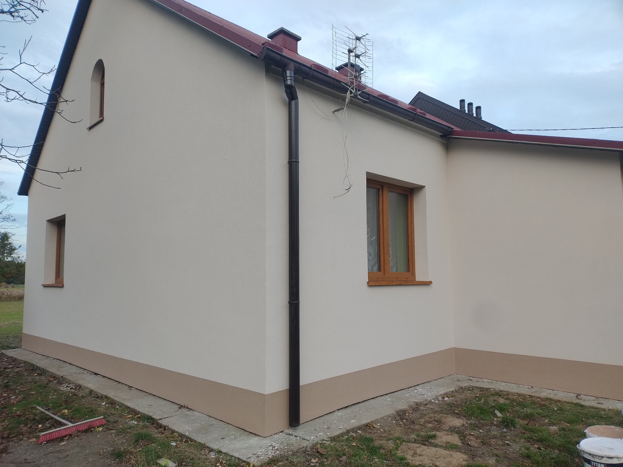 Jasna elewacja domu jednorodzinnego z brązowym cokołem i drewnianą stolarką okienną. Widoczny fragment dachu, rynna spustowa i antena telewizyjna.