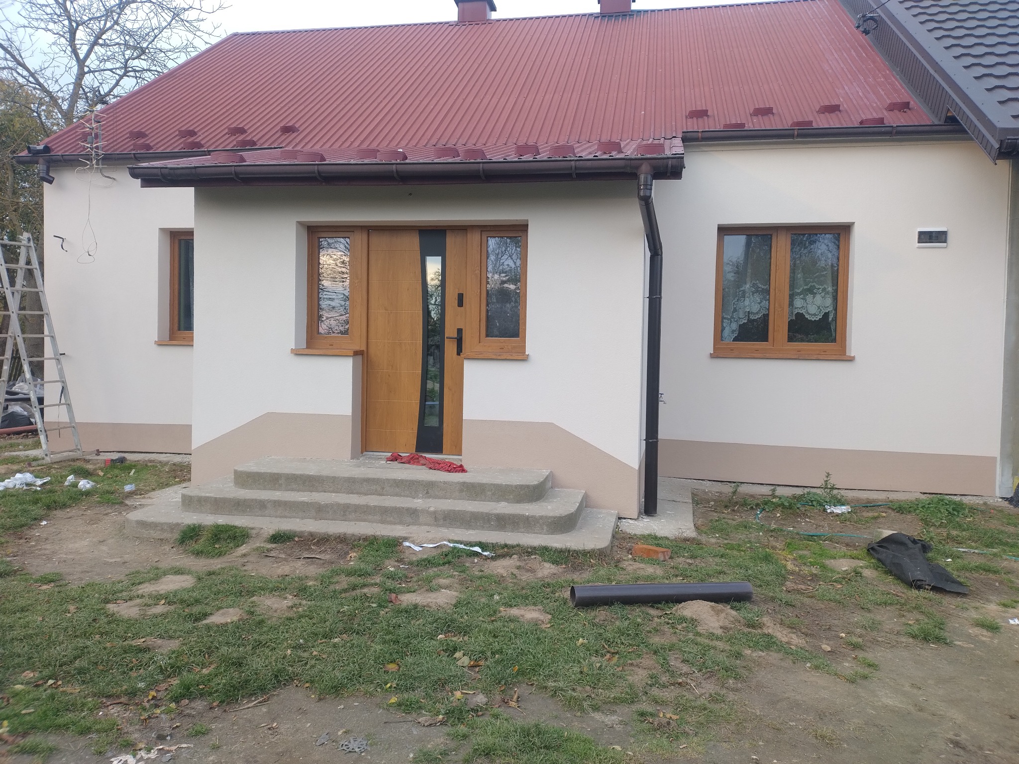 Wykończony dom z nową elewacją w odcieniach bieli i beżu, z brązowymi oknami i drzwiami. Widoczne schody wejściowe i fragment dachu z czerwoną blachodachówką.