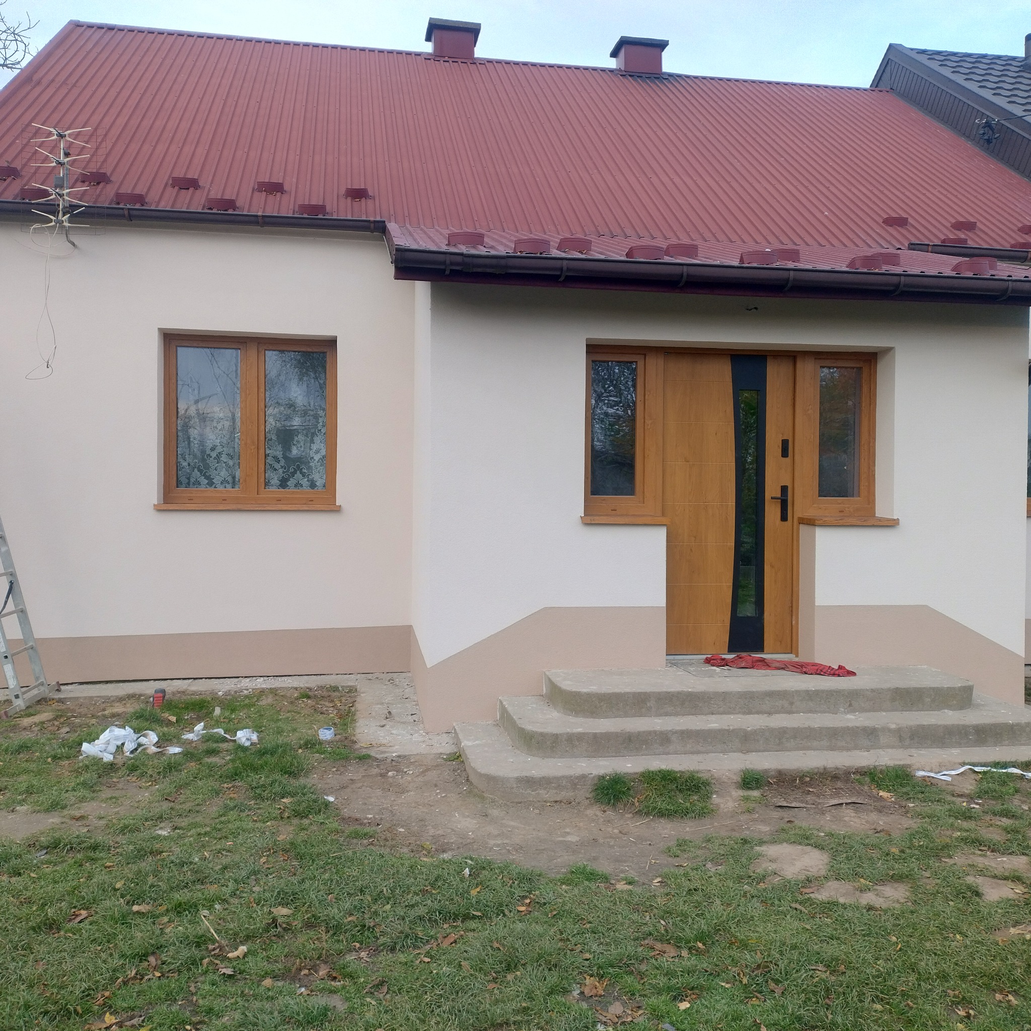 Dom po remoncie elewacji w Radgoszczu. Jasna fasada z drewnianymi oknami i drzwiami. Czerwony dach i betonowe schody wejściowe. Widoczne ślady po pracach.
