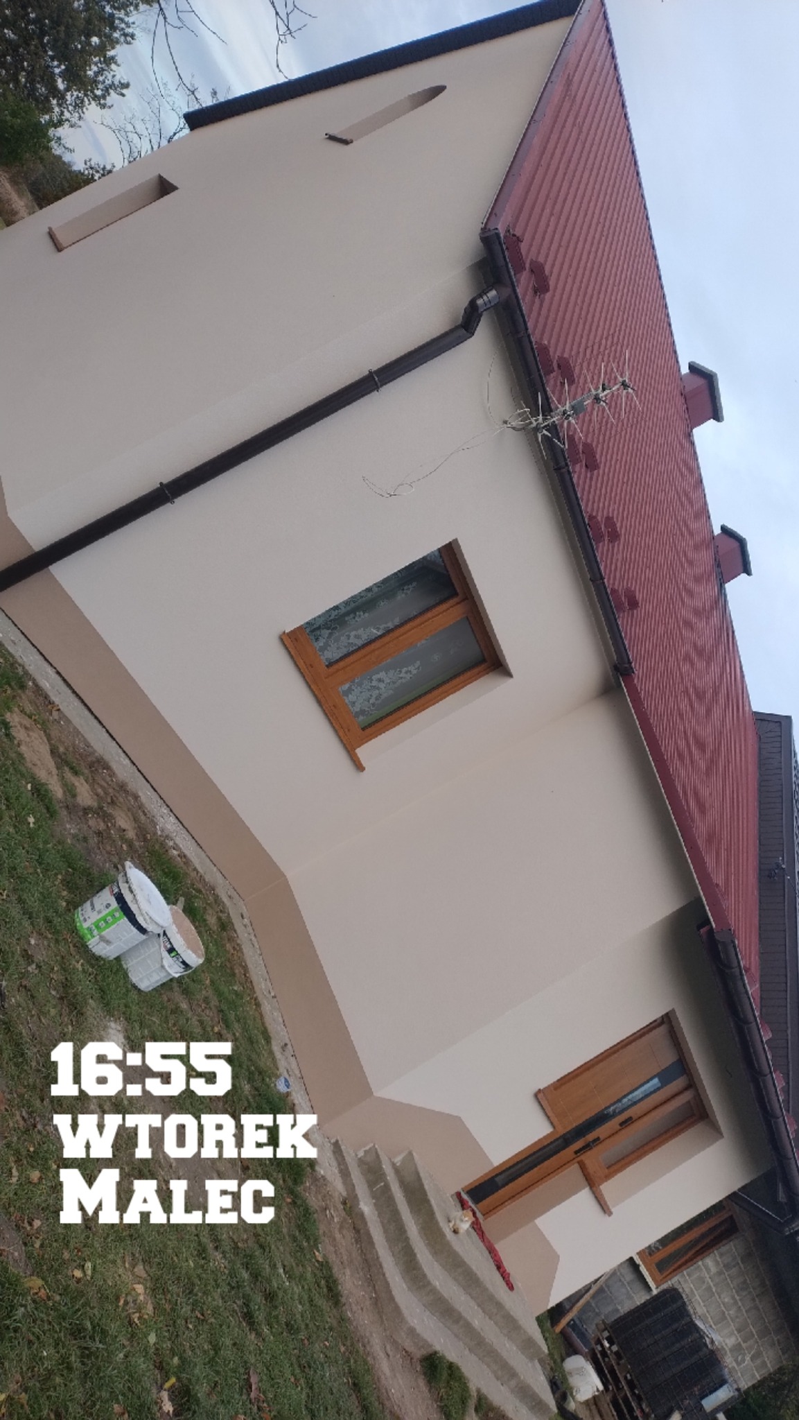 Elewacja domu po malowaniu w dwóch odcieniach beżu, z widocznymi detalami jak okna z drewnianą ramą i brązowe rynny. Na pierwszym planie puszki z farbą.