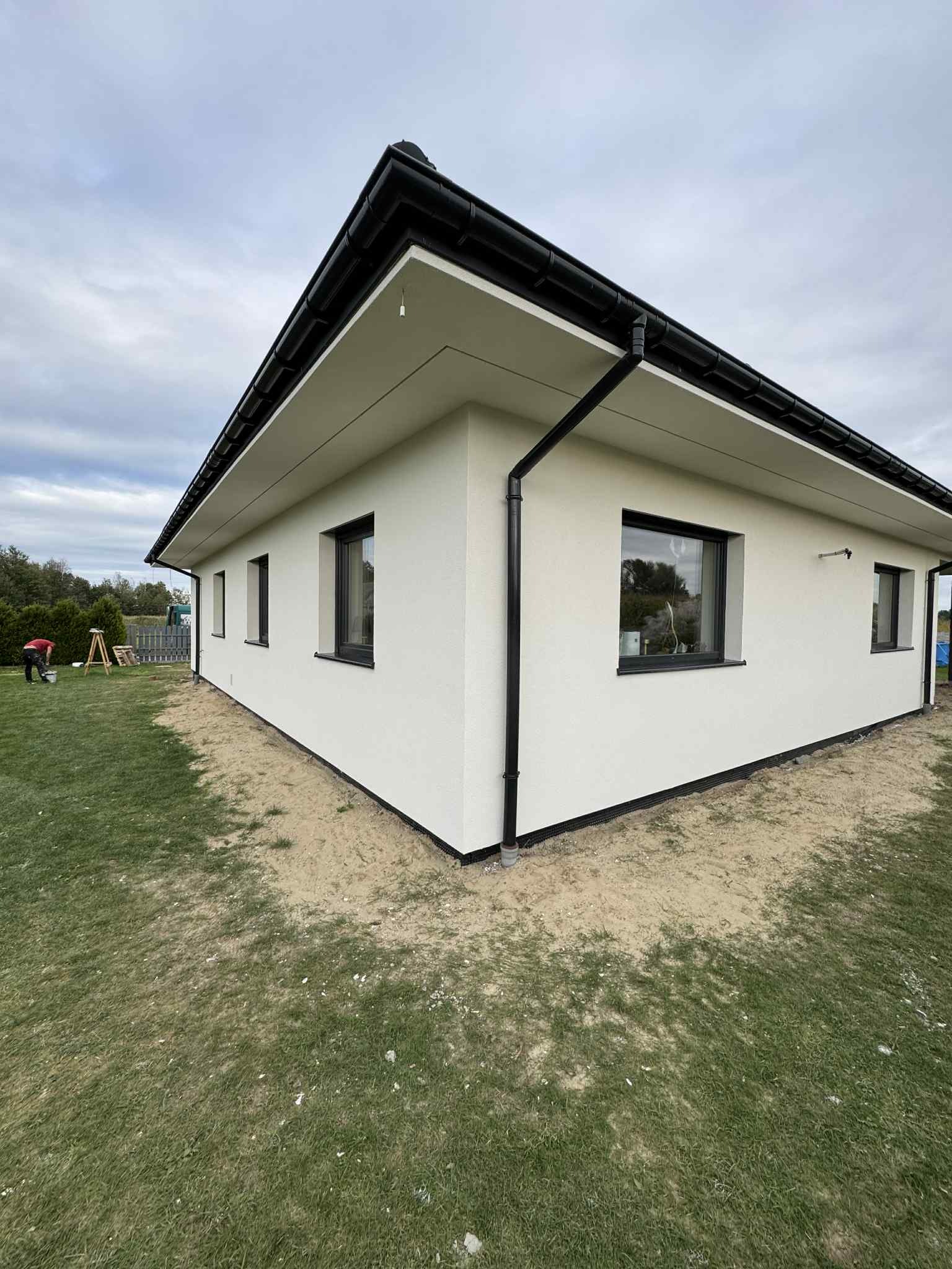 Nowoczesny, parterowy dom z jasną elewacją i czarnymi ramami okiennymi oraz rynnami. Widoczny fragment trawnika i osoba pracująca w oddali.