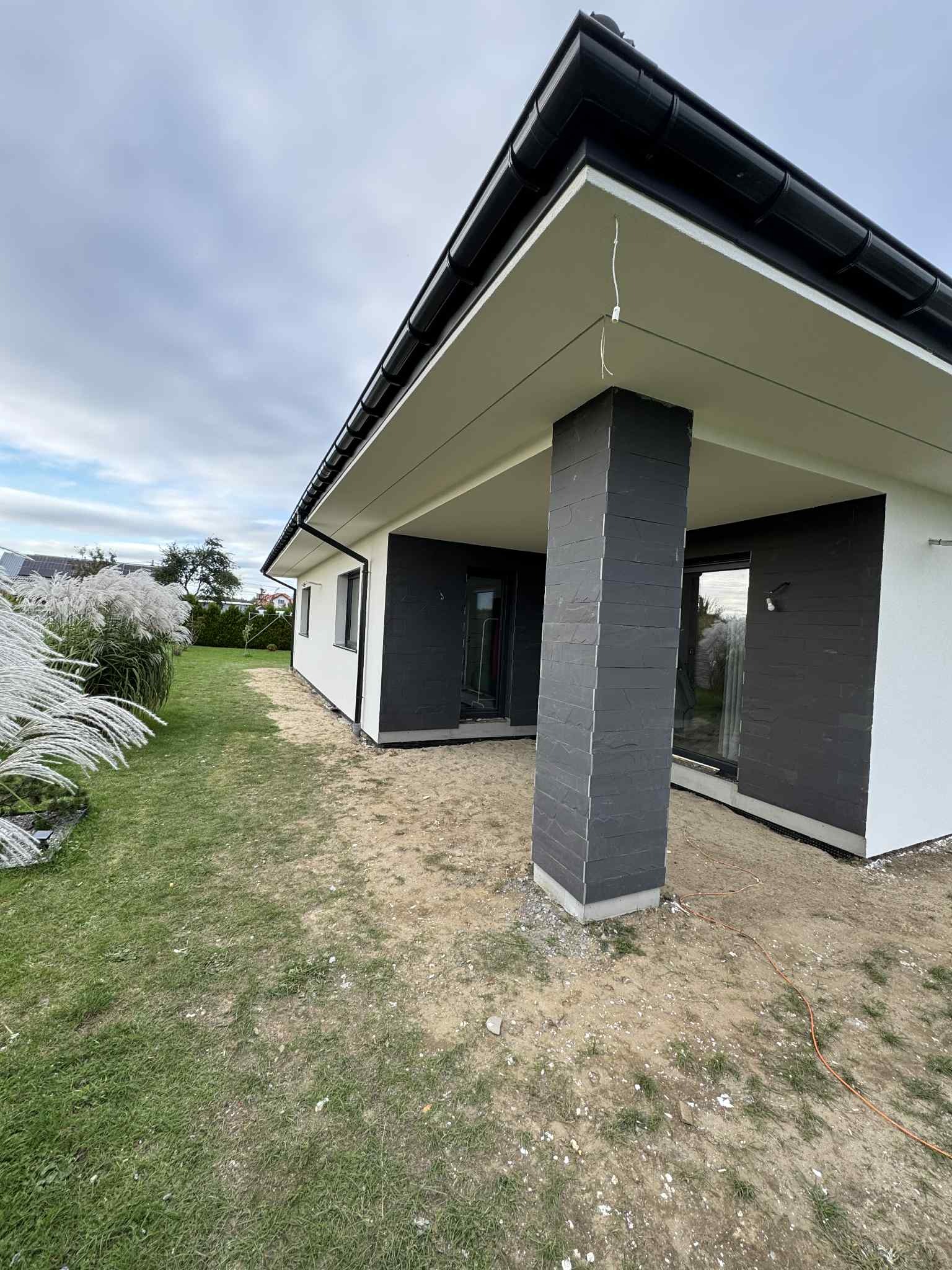 Nowoczesny dom z elewacją w kolorze bieli i grafitu, z kolumną obłożoną kamieniem łupkowym. Widoczne niedokończone prace ziemne wokół budynku.