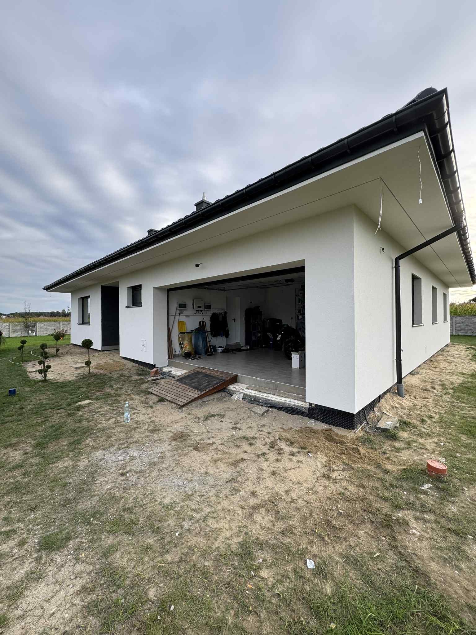 Nowoczesny dom jednorodzinny z białymi ścianami i czarnym dachem, widok z zewnątrz na otwarty garaż. Teren wokół domu w trakcie zagospodarowywania.