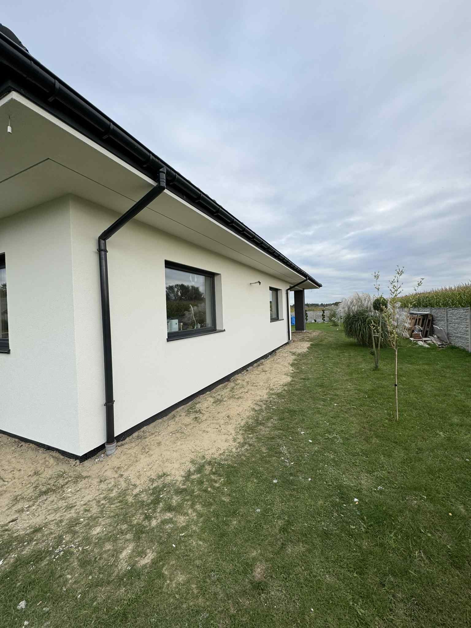 Boczny widok domu z jasną elewacją i czarnymi rynnami, otoczonego trawnikiem i roślinnością. Widoczne okna z czarnymi ramami i fragment ogrodzenia w tle.