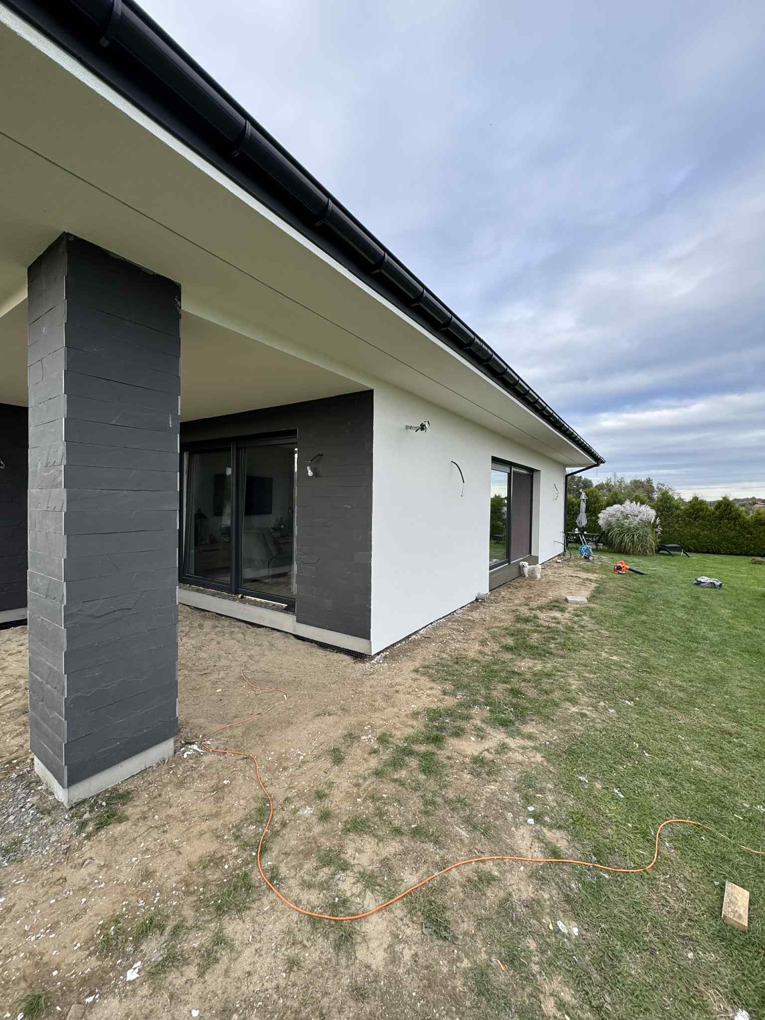 Nowoczesny dom jednorodzinny z wykończoną elewacją: biały tynk i szara okładzina kamienna na filarach i fragmentach ścian. Widoczne duże okna przesuwne i zadbany ogród.