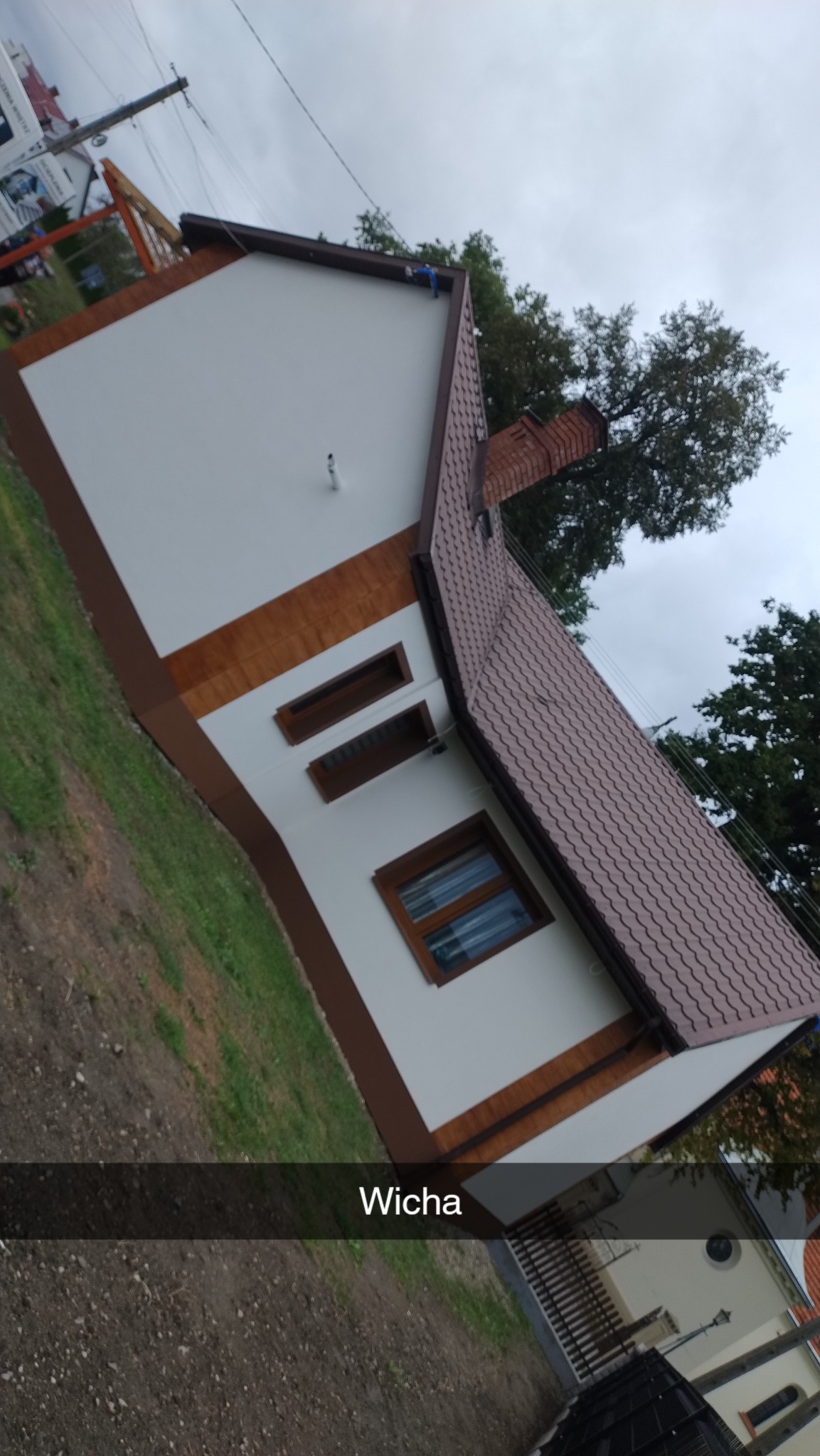 Wykończony dom z białą elewacją, brązowymi akcentami i czerwoną dachówką, widziany z boku pod kątem. Widoczne okna i komin. Pochmurne niebo w tle.