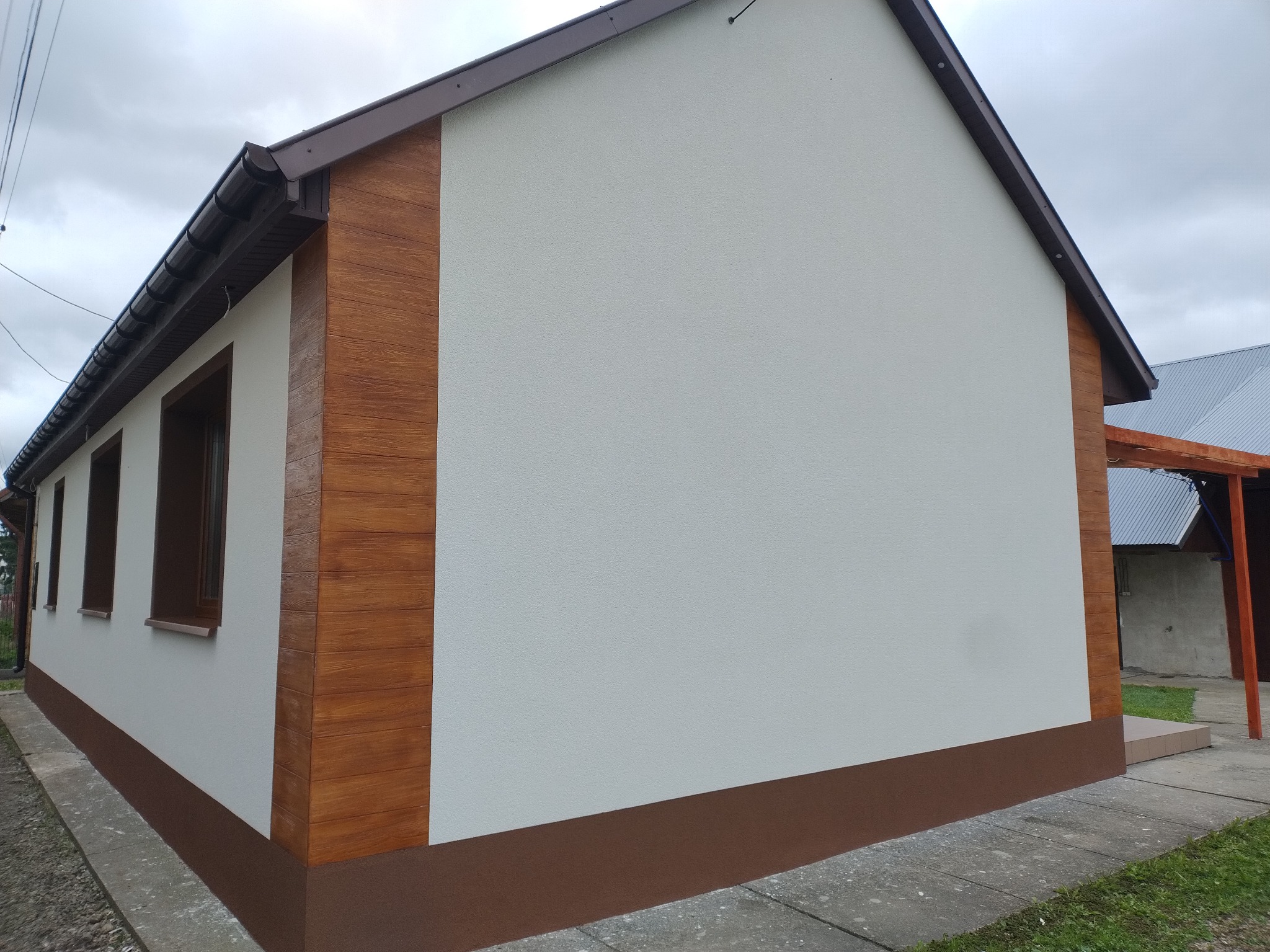 Wykończona elewacja domu z elementami dekoracyjnymi imitującymi drewno, brązowy cokół i orynnowanie. Ściany w jasnym odcieniu, widoczne okna i fragment chodnika.