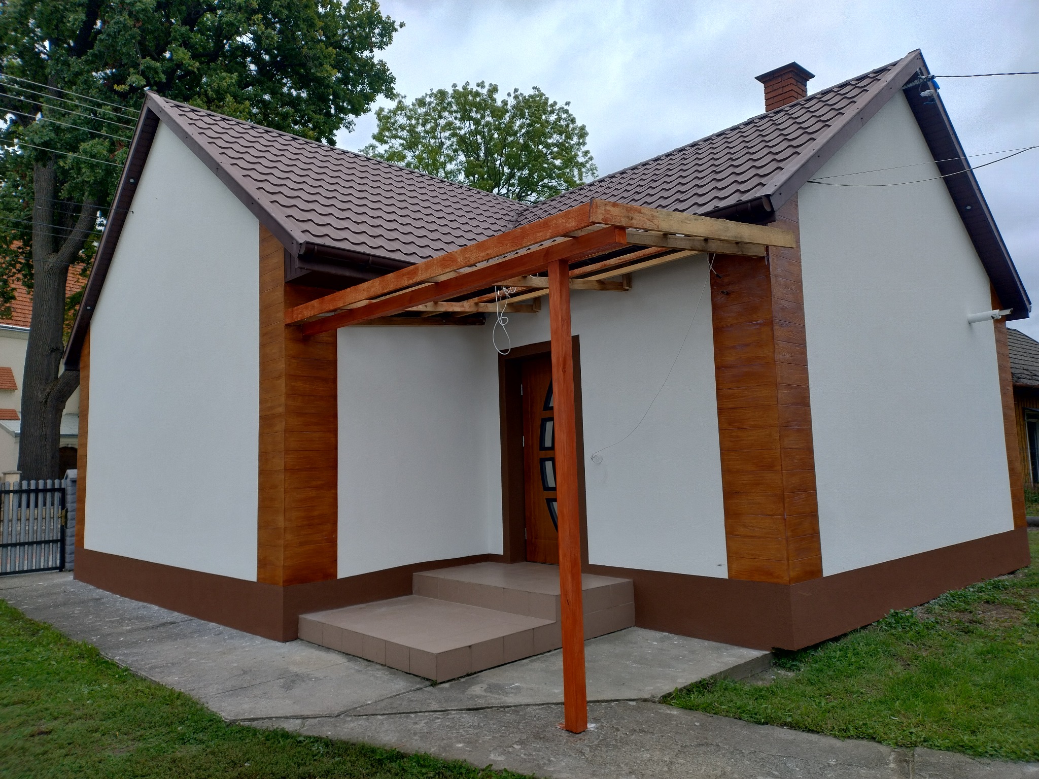 Wykończona elewacja domu z brązowym dachem, drewnianą pergolą nad wejściem i dekoracyjnymi panelami imitującymi drewno. Widoczne schody wejściowe i fragment chodnika.