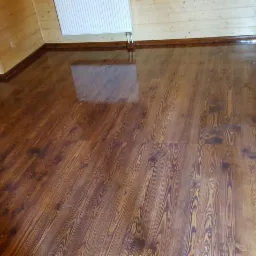 Błyszcząca, brązowa podłoga z desek w drewnianym pokoju z widocznym grzejnikiem i listwami przypodłogowymi.