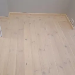 Jasna, odnowiona podłoga z desek, widoczne słoje drewna, listwy przypodłogowe i fragment ściany z gniazdkiem elektrycznym.