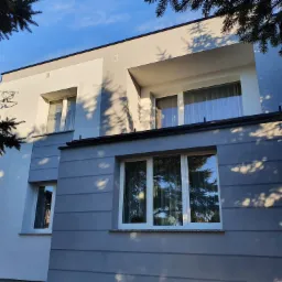 Dwukondygnacyjny dom z szarą elewacją, z poziomymi paskami, białe okna i szara góra, fragment iglastego drzewa w rogu kadru.