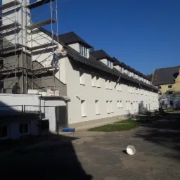 Budynek wielorodzinny w trakcie prac elewacyjnych: ocieplanie i malowanie, widoczne rusztowania i pracownicy w kaskach, częściowo pomalowana fasada na biało.