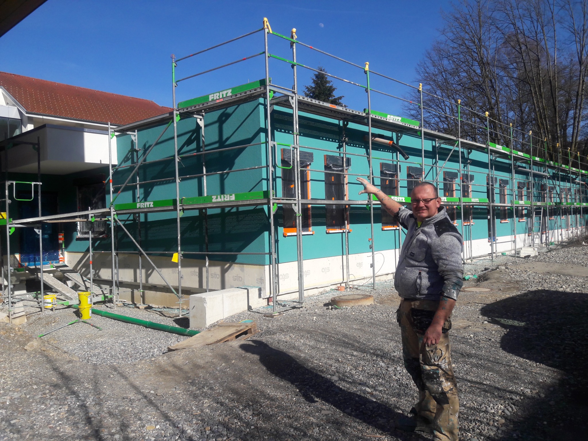 Budynek w trakcie prac elewacyjnych, widoczne rusztowania Fritz, świeżo nałożona warstwa w kolorze turkusowym, pracownik w ubraniu roboczym z wyciągniętą ręką, teren budowy.