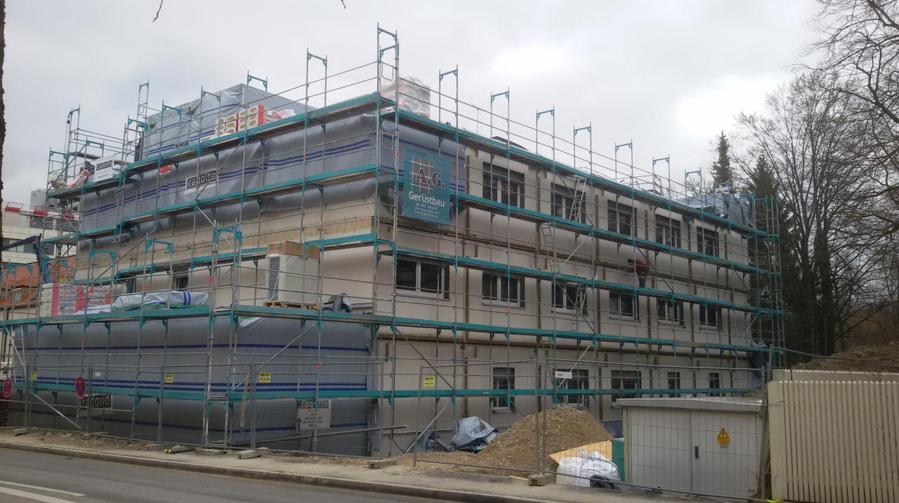 Budynek wielorodzinny w trakcie prac elewacyjnych, otoczony rusztowaniami i częściowo osłonięty folią ochronną z widocznym logo firmy Gerüstbau.