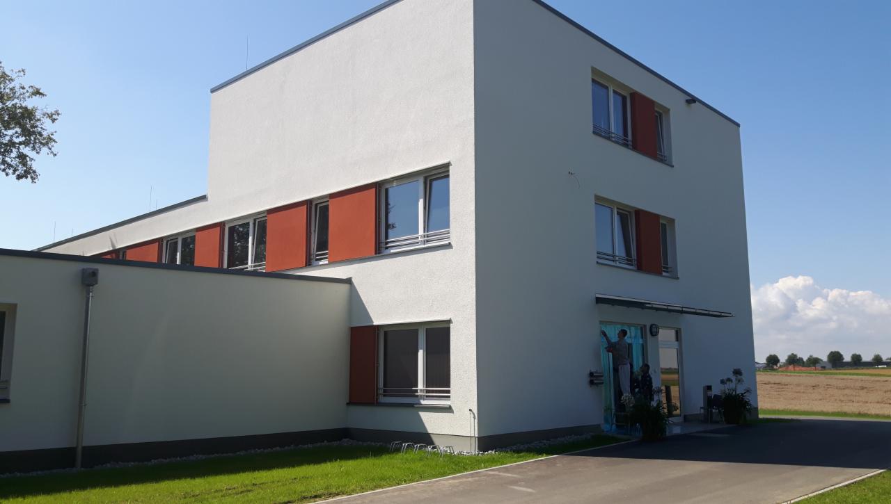 Nowoczesny budynek z białą elewacją i brązowymi okiennicami, widok z zewnątrz na tle błękitnego nieba i pola.