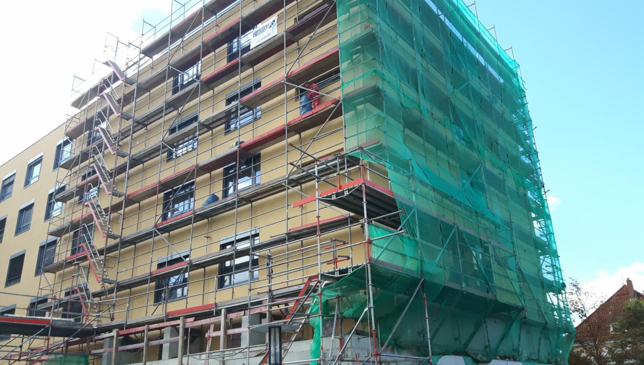 Żółty budynek wielopiętrowy w trakcie prac budowlanych, osłonięty rusztowaniami i zieloną siatką ochronną, z widocznymi pracownikami na rusztowaniach.