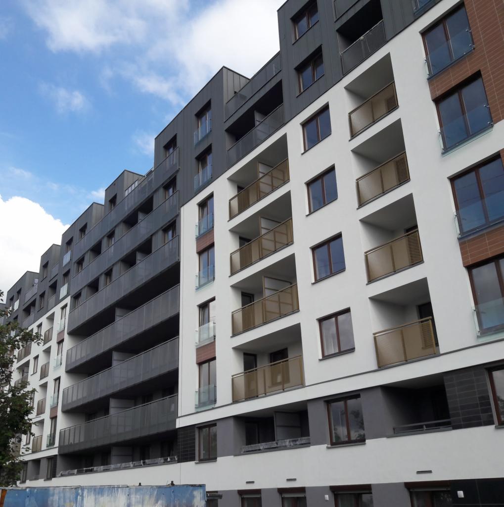 Nowoczesny budynek wielorodzinny w Bielanach Wrocławskich z białymi i grafitowymi elewacjami oraz balkonami z jasnobrązowymi barierkami, sfotografowany w słoneczny dzień z widocznym fragmentem...