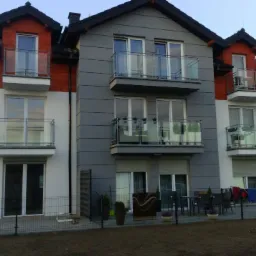 Trzykondygnacyjny budynek wielorodzinny z balkonami, pomalowany w odcieniach szarości, bieli i czerwieni, z widocznymi elementami wykończeniowymi elewacji i zagospodarowania terenu wokół budynku.