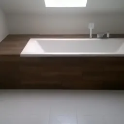 Biała wanna w obudowie z płytek imitujących drewno, w jasnej łazience z białymi płytkami podłogowymi i oknem dachowym.