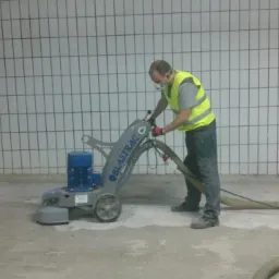 Pracownik w kamizelce i masce szlifuje betonową posadzkę za pomocą maszyny Blastraco w pomieszczeniu z wyłożonymi kafelkami ścianami. Widoczny wąż odkurzacza.