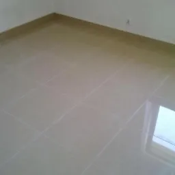 Jasne, błyszczące płytki podłogowe w narożniku pokoju, z widoczną listwą przypodłogową i odbiciem okna. Minimalistyczne wykończenie, neutralna kolorystyka.