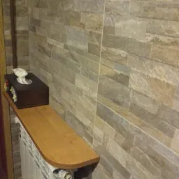 Wykończona łazienka z kamienną okładziną ściany, drewnianą półką nad grzejnikiem i ozdobnym pudełkiem na chusteczki. Detal wykończenia wnętrza.