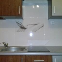 Rajmund Kamiński - Wykończona kuchnia z białymi płytkami z motywem sztućców nad zlewem i płytą grzewczą. Widoczne szafki wiszące i blat.